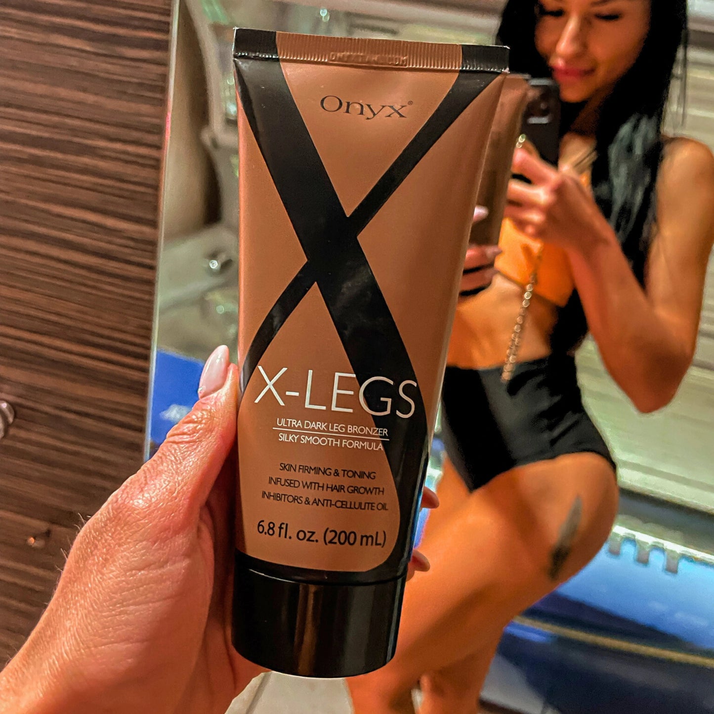 Zbliżenie na tubkę Onyx X-LEGS przed lustrem, kobieta w tle ubrana w strój do opalania